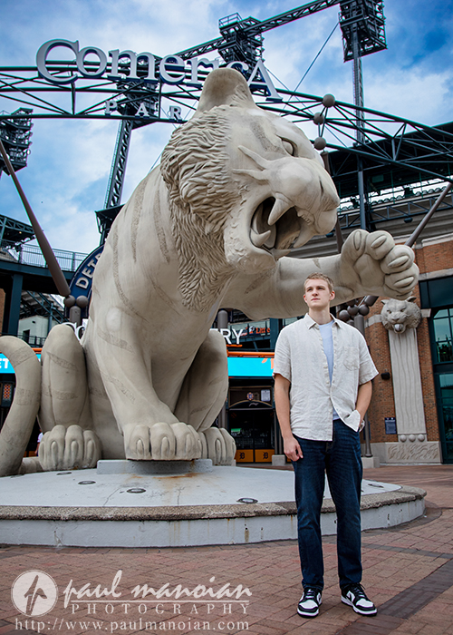 A high school senior boy stands in front of a large roaring tiger statue at Comerica Park, baseball stadium in Detroit, Michigan, during a session for White Lake senior pictures under a partly cloudy sky.