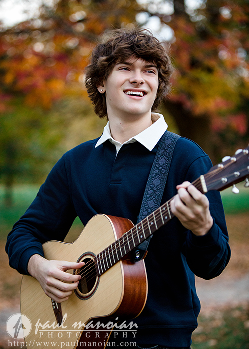A young man with wavy brown hair smiles while playing an acoustic guitar outdoors in a park during his Birmingham senior pictures session, surrounded by autumn trees with colorful leaves. He wears a navy sweater over a white collared shirt.