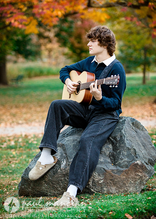 A young person with curly hair sits on a large rock in a park during autumn, playing an acoustic guitar. Colorful fall leaves and trees create the perfect backdrop for a memorable Birmingham senior pictures session.
