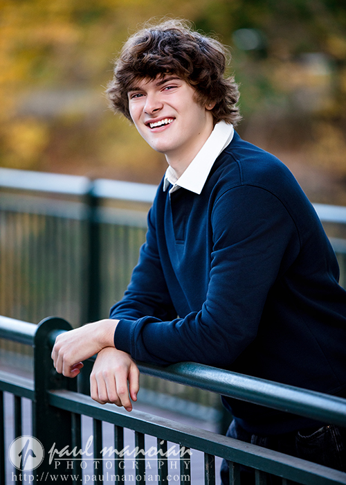 A young person with curly brown hair, wearing a dark blue sweater over a white collared shirt, smiles while leaning on a green metal railing outdoors during their Birmingham senior pictures session. Trees with autumn colors are blurred in the background.