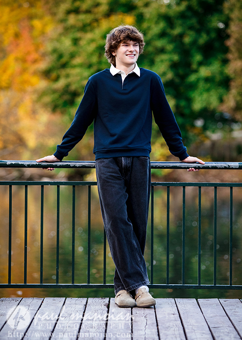 A young person with curly brown hair, wearing a navy sweater and dark jeans, leans casually on a black railing in front of a lake with autumn-colored trees—a perfect moment from a Birmingham senior pictures session.
