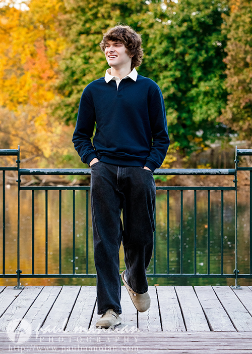 A young man with curly brown hair walks on a wooden dock, smiling, during his Birmingham senior pictures session. He wears a navy sweater, white collared shirt, and dark jeans. Autumn trees and water create a picturesque backdrop.