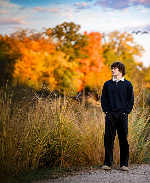 A young man stands on a gravel path surrounded by tall grass, with colorful autumn trees and a flock of birds flying in the cloudy sky behind him. Captured during a Birmingham senior pictures session, he looks to the right, hands in his pockets, wearing dark clothes.