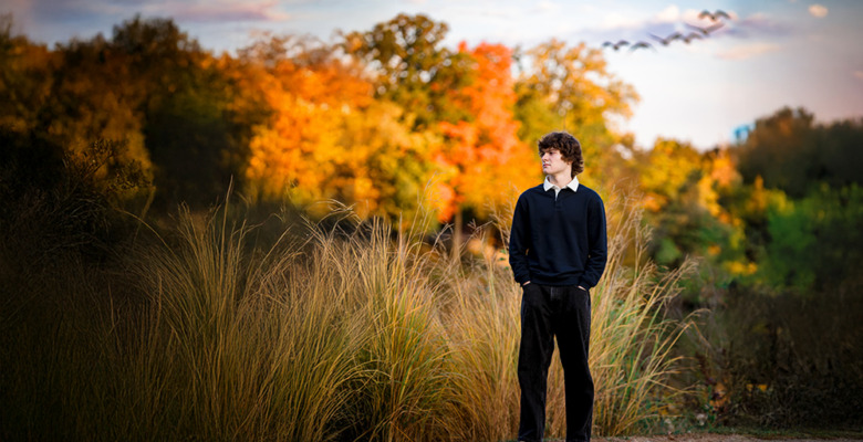A young man stands on a gravel path surrounded by tall grass, with colorful autumn trees and a flock of birds flying in the cloudy sky behind him. Captured during a Birmingham senior pictures session, he looks to the right, hands in his pockets, wearing dark clothes.