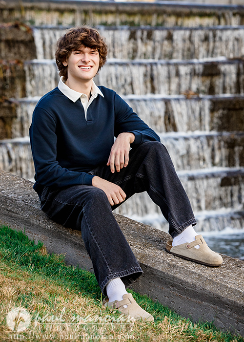 A young person with wavy brown hair, wearing a navy sweater, dark jeans, and tan shoes, sits on a concrete edge near tiered waterfalls during a Birmingham senior pictures session, smiling at the camera. Grass is visible in the foreground.