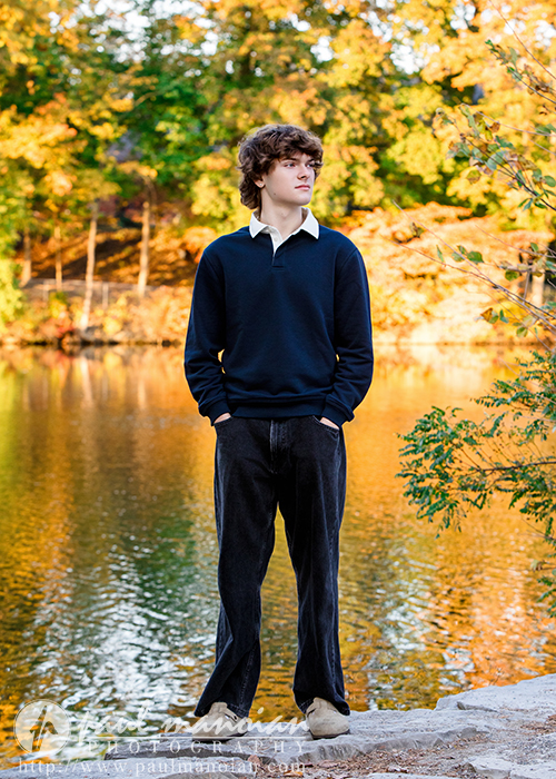 A young person with curly brown hair stands by a lake during their Birmingham senior pictures session, wearing a navy blue sweater and collared shirt. Autumn trees with golden leaves are reflected in the water behind them.