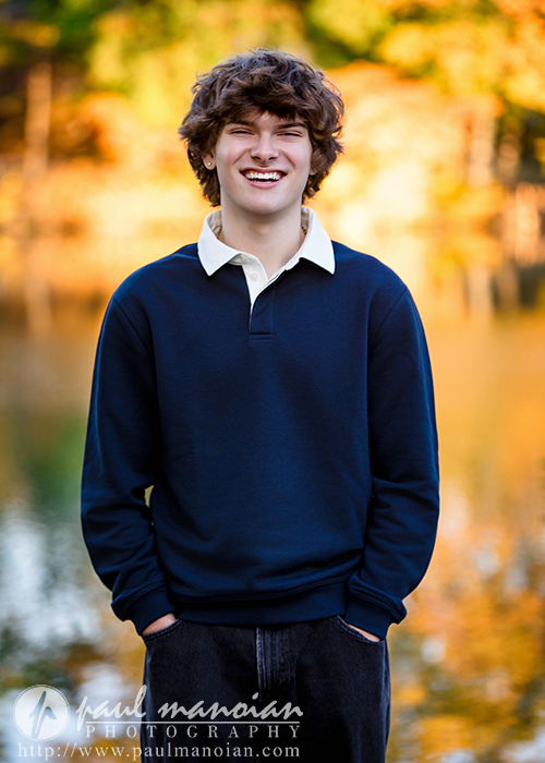 A young man with curly brown hair, wearing a navy sweater over a white collared shirt, stands outdoors smiling with his hands in his pockets during his Birmingham senior pictures session. Autumn trees and a blurred lake are in the background.