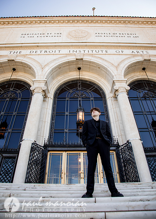 A person in a black suit stands on the steps outside the Detroit Institute of Arts, with tall arched doorways and columns in the background under a clear sky.