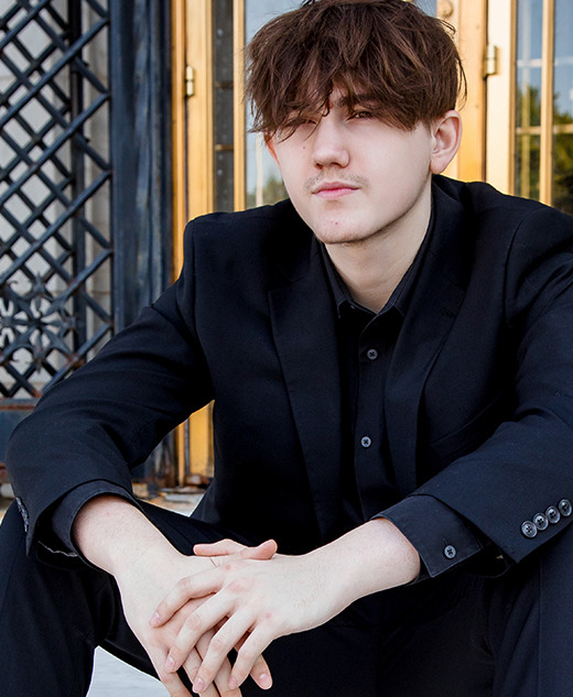 A young man in a black suit sits on marble steps with his hands clasped, in front of ornate iron and glass doors. His hair is slightly messy and he gazes forward with a neutral expression.