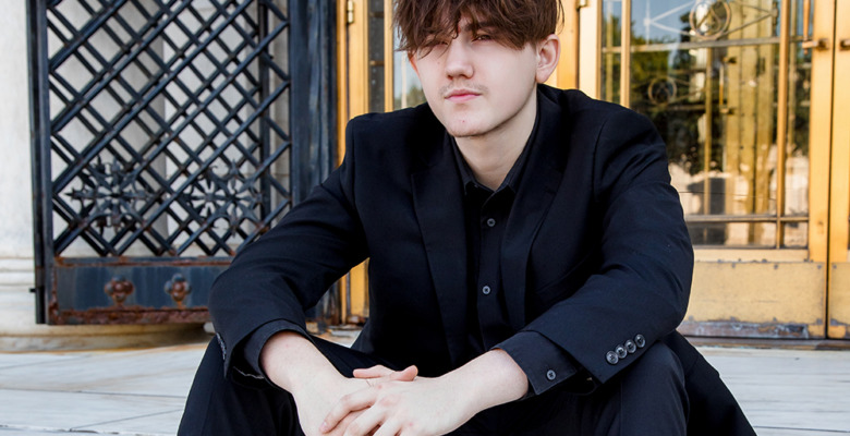 A young man in a black suit sits on marble steps with his hands clasped, in front of ornate iron and glass doors. His hair is slightly messy and he gazes forward with a neutral expression.