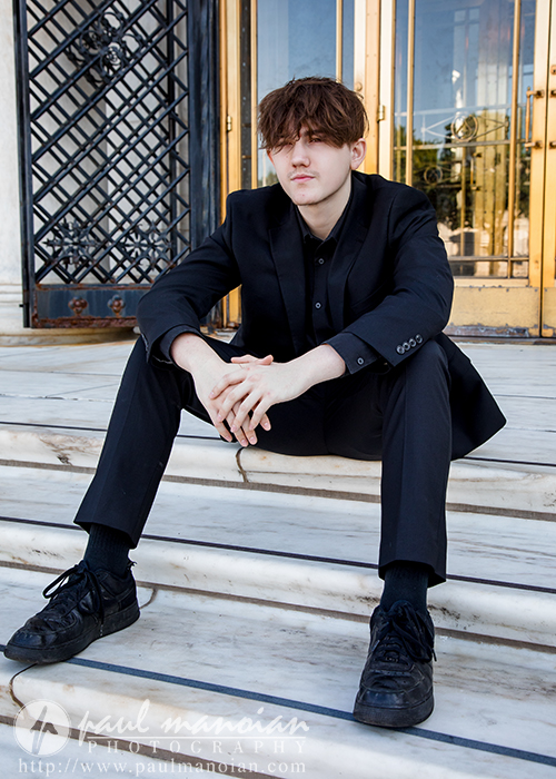 A young man in a black suit and black sneakers sits on marble steps outside a building with ornate metal doors, looking relaxed with his arms resting on his knees.