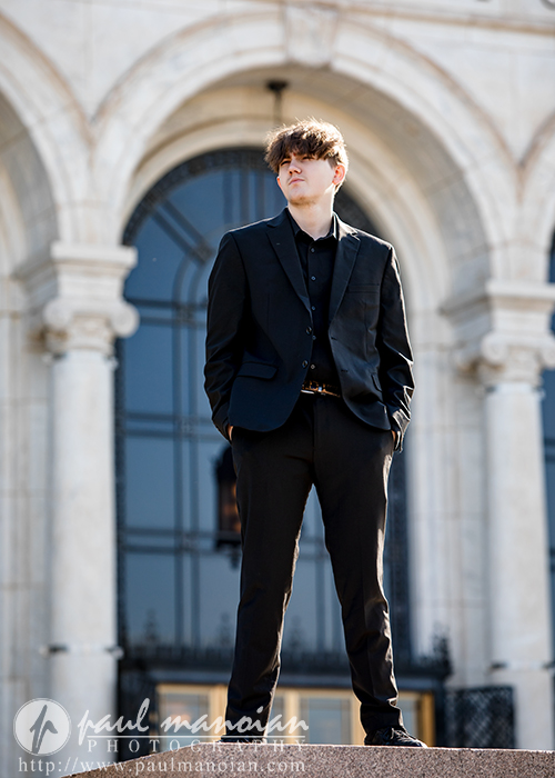 A young man in a black suit stands confidently outside a building with large arched windows and ornate columns, looking off to the side.