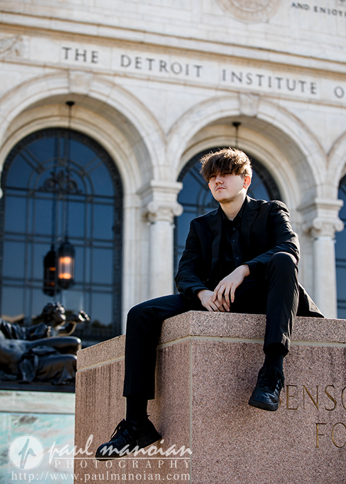 A young person in a black suit sits on a stone pedestal in front of The Detroit Institute of Arts, with arched windows and a sculpture visible in the background.