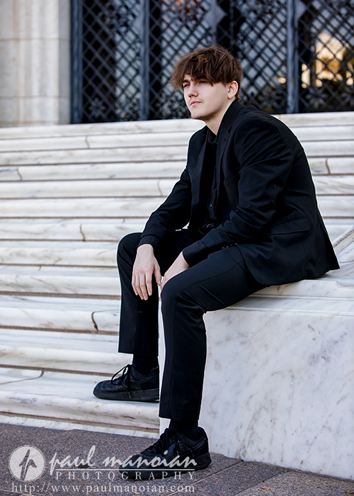 A young man in a black suit sits on marble steps outside a building with black, decorative metal doors. He looks off to the side with a neutral expression. The image is credited to Paul Manoian Photography.
