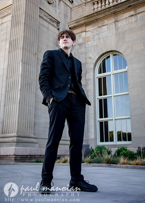 A young man wearing a black suit stands confidently outside a large stone building with tall windows and columns. The photo is taken from a low angle, emphasizing the building’s architectural details.