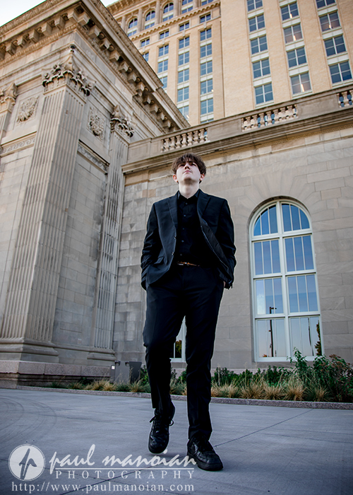 A young man in a black suit stands confidently in front of a tall, historic stone building with large windows and columns, looking slightly upward. The sky is clear and the setting is urban.
