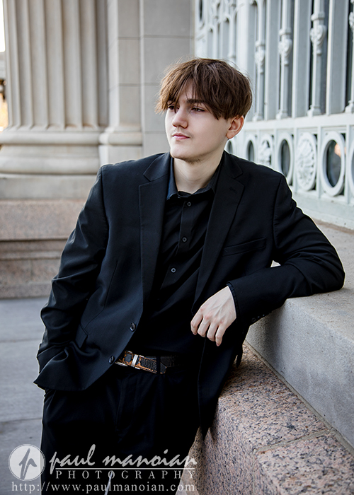 A young man in a black suit leans casually against a stone railing outdoors, with one hand in his pocket. Ornate metal and stone architectural details are visible in the background.