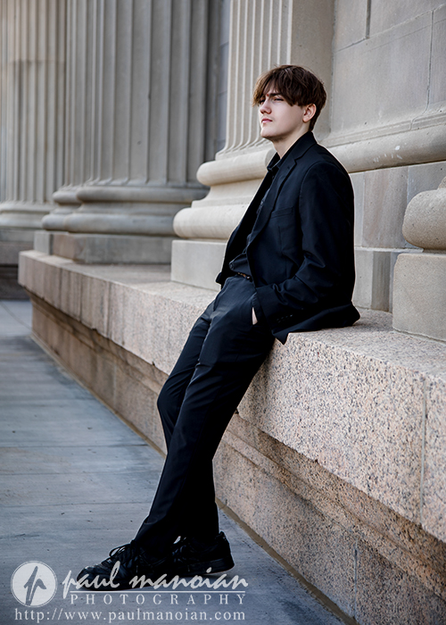A young man in a black suit leans casually against a stone ledge in front of a building with large columns, looking off to the side with his hands in his pockets.