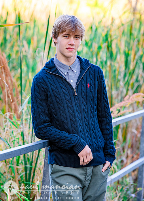 A young man wearing a navy blue cable-knit sweater and khaki pants leans casually against a metal railing outdoors, with tall green and yellow grasses blurred in the background.