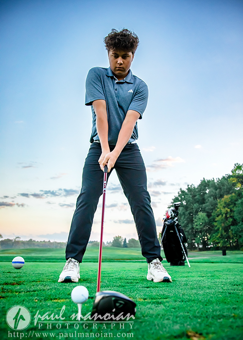 A young golfer stands ready to swing, focusing on a golf ball on a tee at a green course. Golf clubs rest in a bag nearby, with trees and a cloudy sky in the background.