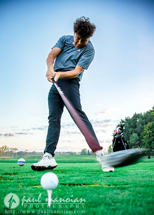 A young golfer in mid-swing hits a golf ball on a green field at sunrise or sunset, with golf tees and a golf bag visible in the background. The sky is partly cloudy, and the grass is vibrant green.