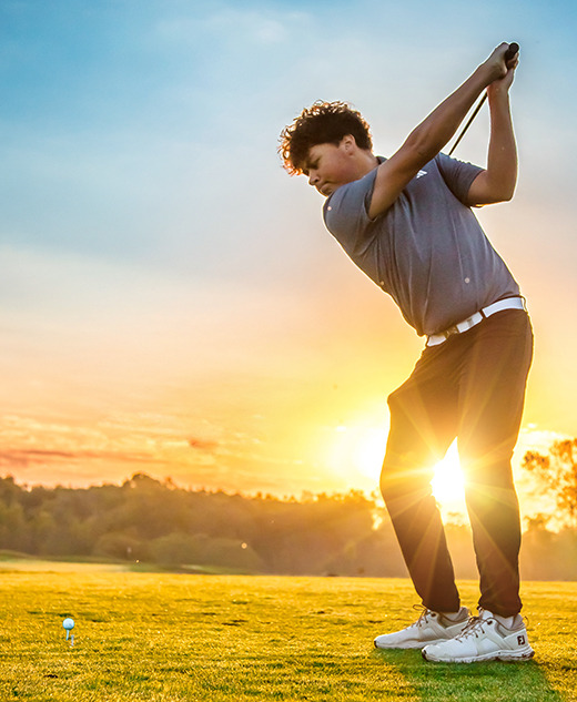 A high school senior boy prepares to swing a golf club at a golf ball on a grassy course during a stunning sunset, trees in the background—an ideal moment for a golf senior pictures session.
