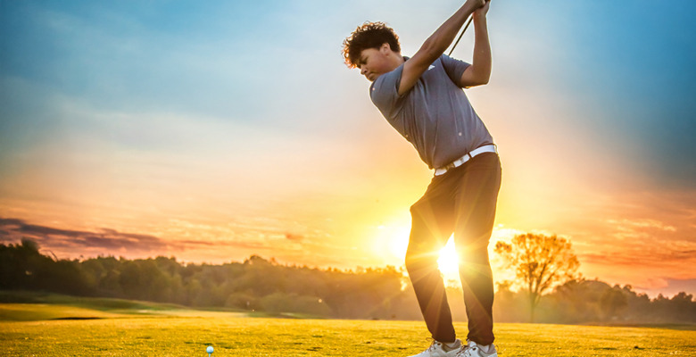 A high school senior boy prepares to swing a golf club at a golf ball on a grassy course during a stunning sunset, trees in the background—an ideal moment for a golf senior pictures session.