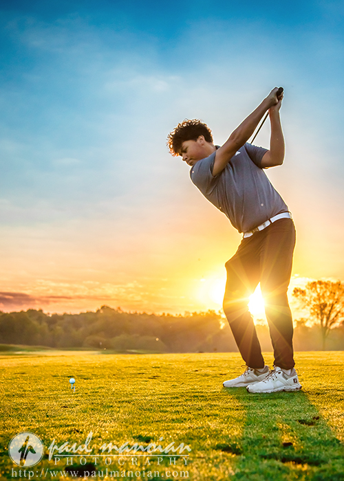 A young man prepares to swing a golf club on a grassy course at sunset, with warm sunlight shining behind him and trees visible in the background.