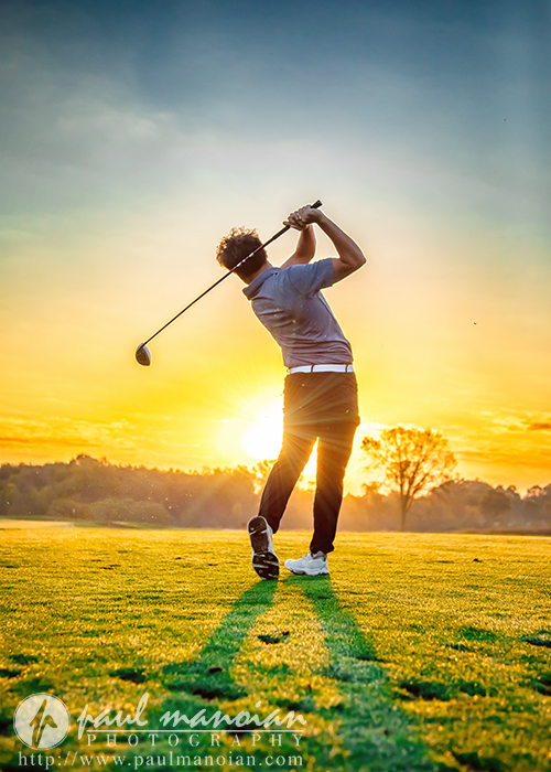 A golfer swings a club on a grassy course at sunset, casting a long shadow. The sky is vibrant with orange and yellow hues, and trees are visible in the background.