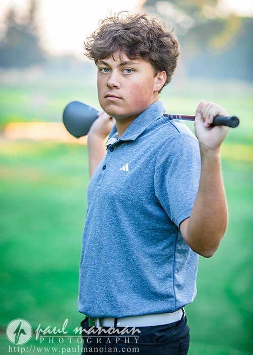 Teen boy in a blue polo shirt stands on a golf course, holding a golf club across his shoulders, looking directly at the camera with a serious expression. The background is blurred greenery.