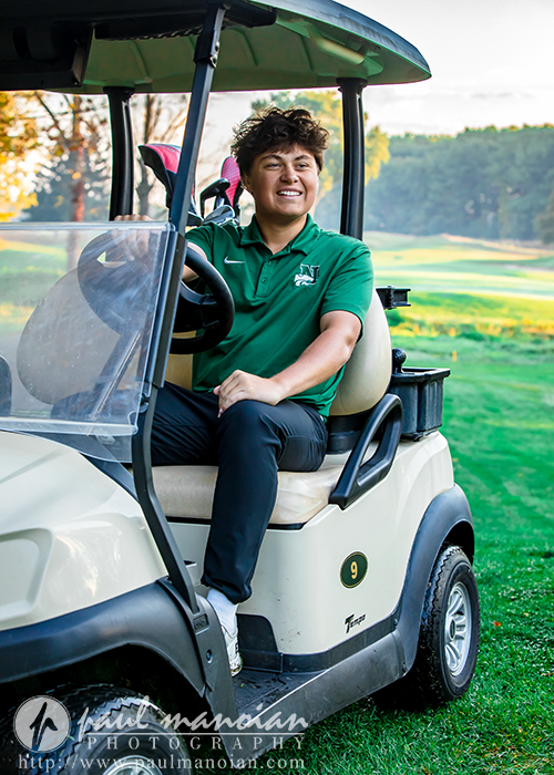A young person in a green polo shirt sits smiling in a golf cart on a golf course, with golf clubs visible in the back and green fairways in the background.