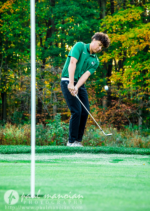 A young golfer in a green shirt and black pants focuses as he chips a golf ball toward the green, with trees and autumn foliage in the background. The flagstick and hole are visible in the foreground.