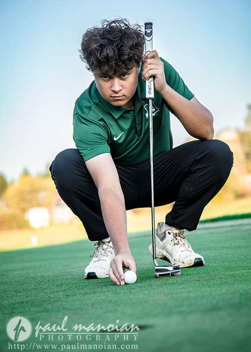A young golfer in a green shirt and black pants crouches on a green, carefully placing a golf ball while holding a putter, with a focused expression. Outdoor golf course setting, logo watermark at the bottom.