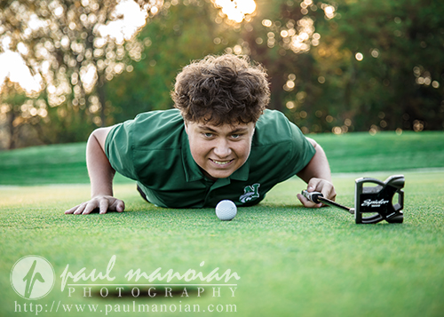 A young person in a green shirt lies on a golf course, lined up with a golf ball and putter, smiling at the camera. Trees and sunlight are in the background.