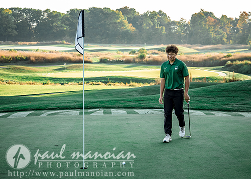 A young golfer in a green shirt and black pants walks on a putting green with a putter in hand. The golf course is lush and scenic, with trees and sunlight in the background. A flagstick is in the foreground.