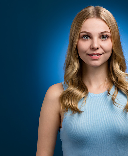 A young woman with long blonde hair and a light blue sleeveless top smiles while standing against a dark blue background with a soft blue halo effect behind her.