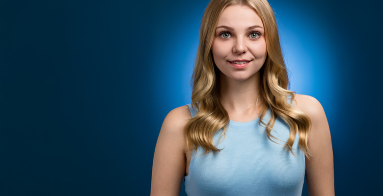A young woman with long blonde hair and a light blue sleeveless top smiles while standing against a dark blue background with a soft blue halo effect behind her.