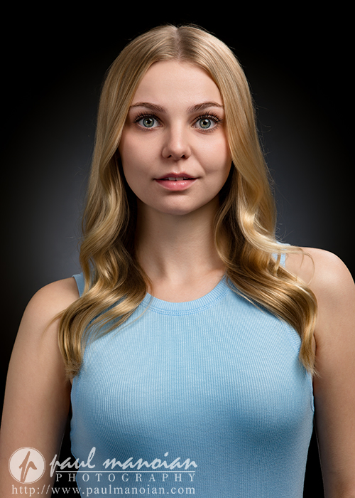 A young woman with long, wavy blonde hair wearing a light blue sleeveless top poses against a dark gradient background during a musical theatre headshot session. She faces forward with a neutral expression.