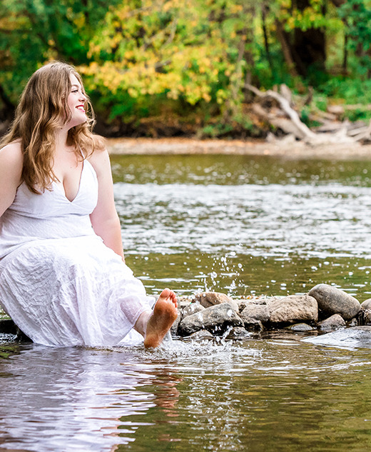 A woman in a white dress sits on rocks by a river, smiling and dipping her bare feet in the water, with green trees and foliage in the background.