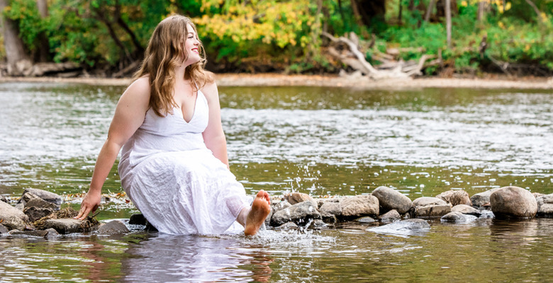 A woman in a white dress sits on rocks by a river, smiling and dipping her bare feet in the water, with green trees and foliage in the background.