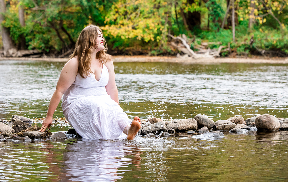 A woman in a white dress sits on rocks by a river, smiling and dipping her bare feet in the water, with green trees and foliage in the background.
