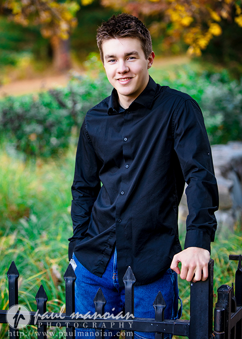 A young man with short brown hair, wearing a black button-up shirt and blue jeans, stands outdoors by a metal fence, smiling during his Ann Arbor senior pictures session, with green grass and trees in the background.