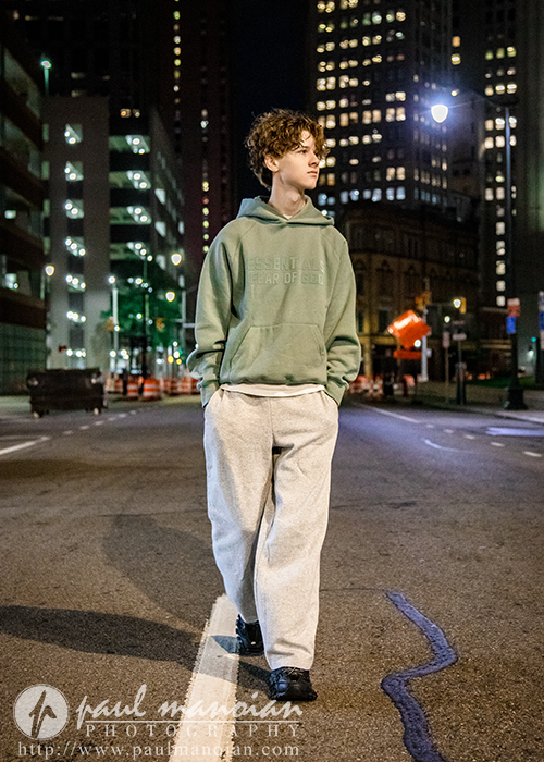 A young person with curly hair, wearing a green hoodie and gray pants, walks alone on a city street at night—tall buildings and streetlights setting the scene for a memorable Detroit senior pictures session.