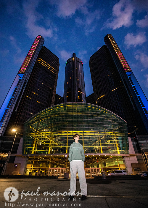 A person in a green hoodie and gray pants stands in front of the illuminated glass entrance of the GM Renaissance Center skyscrapers at dusk, with a dramatic blue and purple sky overhead—perfect for a Detroit senior pictures session.