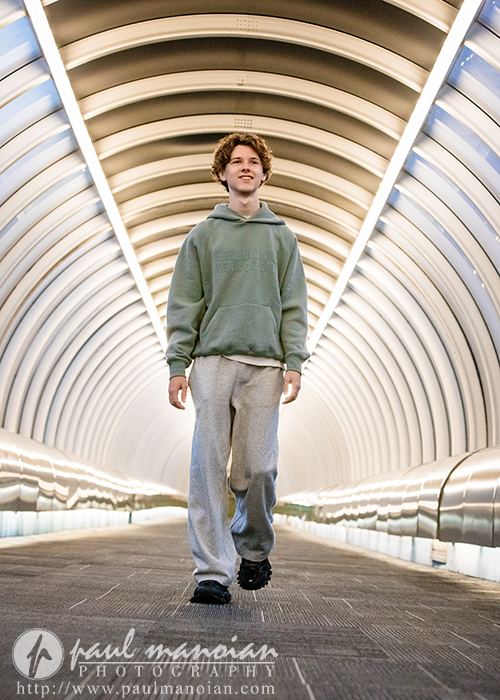 A young person in a green hoodie and gray sweatpants walks confidently through a brightly lit, curved tunnel with modern architectural design, capturing the vibrant energy of a Detroit senior pictures session.