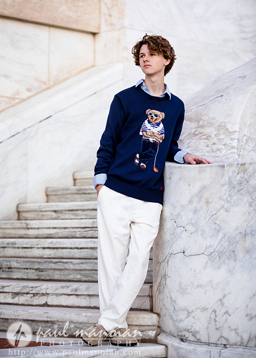 Young man with wavy hair wearing a navy sweater featuring a bear design, light blue shirt, and white pants, leaning against a marble wall next to curved marble steps during a Detroit senior pictures session.