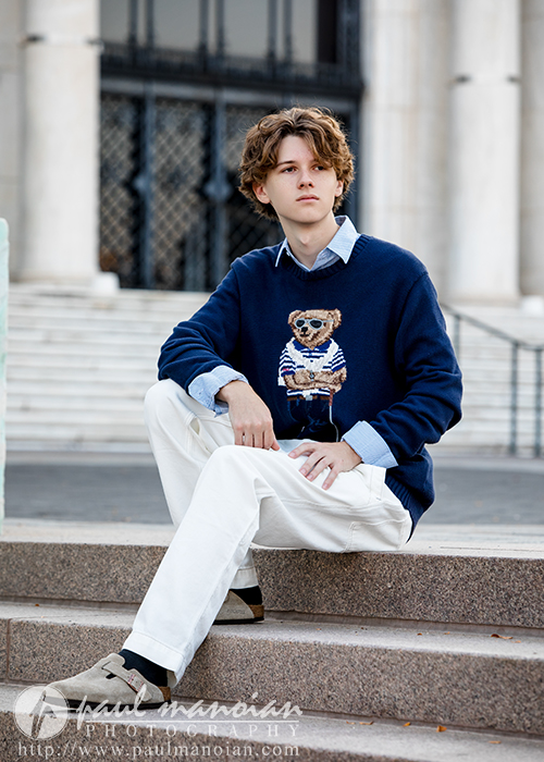 A young person with wavy brown hair sits on outdoor steps during their Detroit senior pictures session, wearing a blue sweater with a bear graphic, white pants, and light shoes. Stone columns and a staircase railing frame the scene.