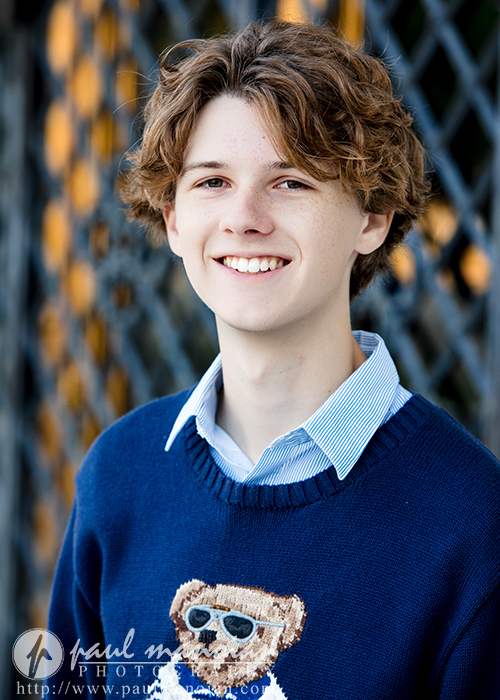 A smiling young person with wavy brown hair wears a blue sweater featuring a bear with sunglasses over a collared shirt, captured during a Detroit senior pictures session. The background is a blurred metal fence with circular patterns.