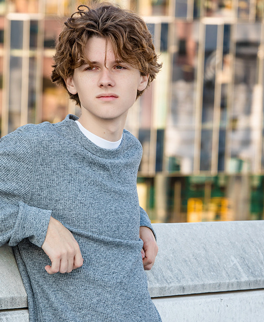 A young person with wavy brown hair, wearing a gray sweater, leans against a concrete wall outdoors during a Detroit senior pictures session, with a modern glass building in the background.