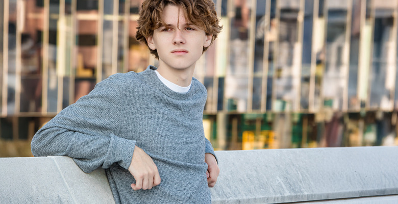 A young person with wavy brown hair, wearing a gray sweater, leans against a concrete wall outdoors during a Detroit senior pictures session, with a modern glass building in the background.